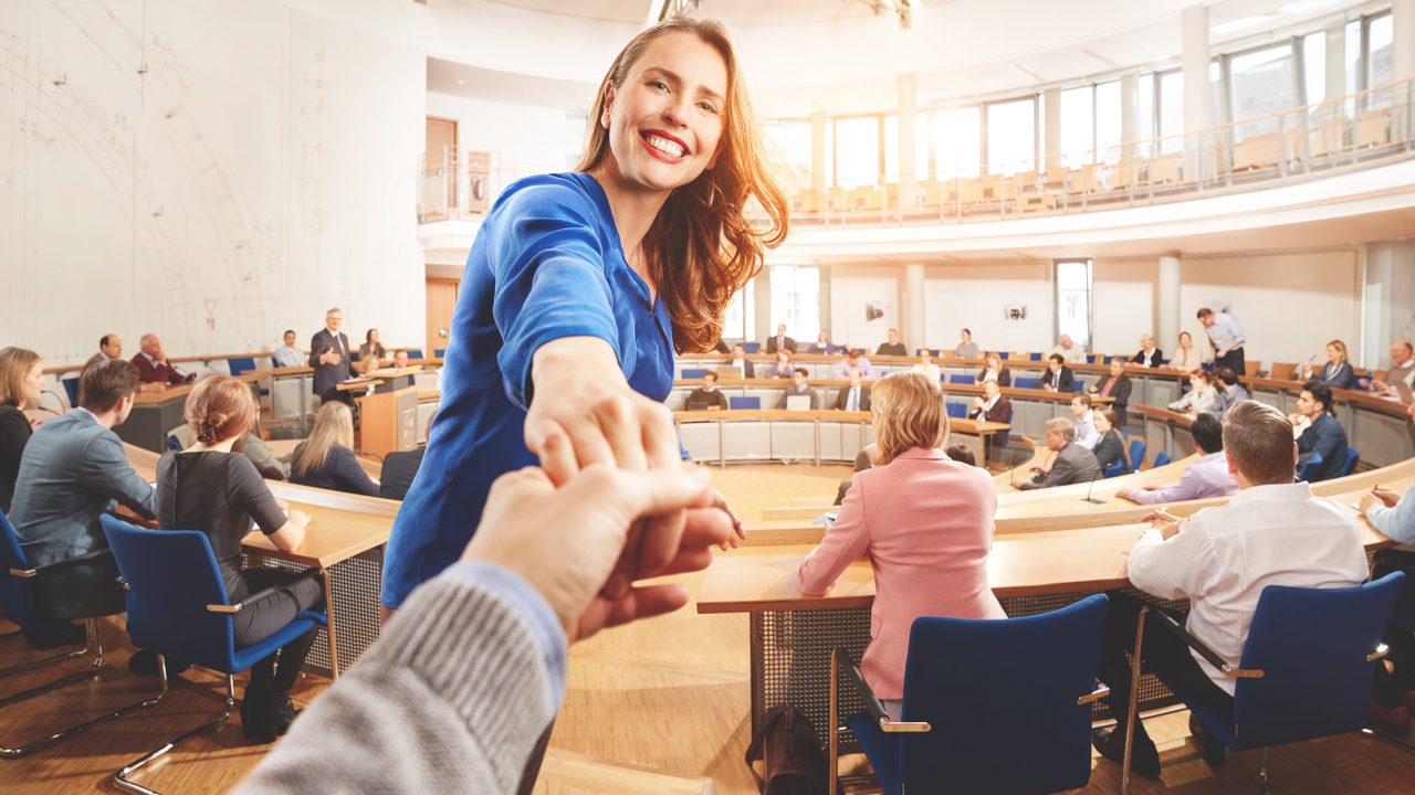Foto: Frau in einem Konferenzsaal reicht jemandem die Hand