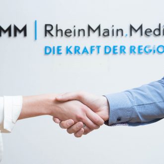 Foto: Zwei schüttelnde Hände vor dem Firmenschild der RheinMain.Media