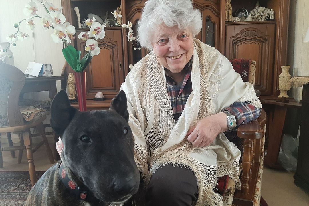 Ältere Seniorin mit weißen Haaren sitzt mit ihrem Hund in ihrem Wohnzimmer und sieht freundlich in die Kamera.