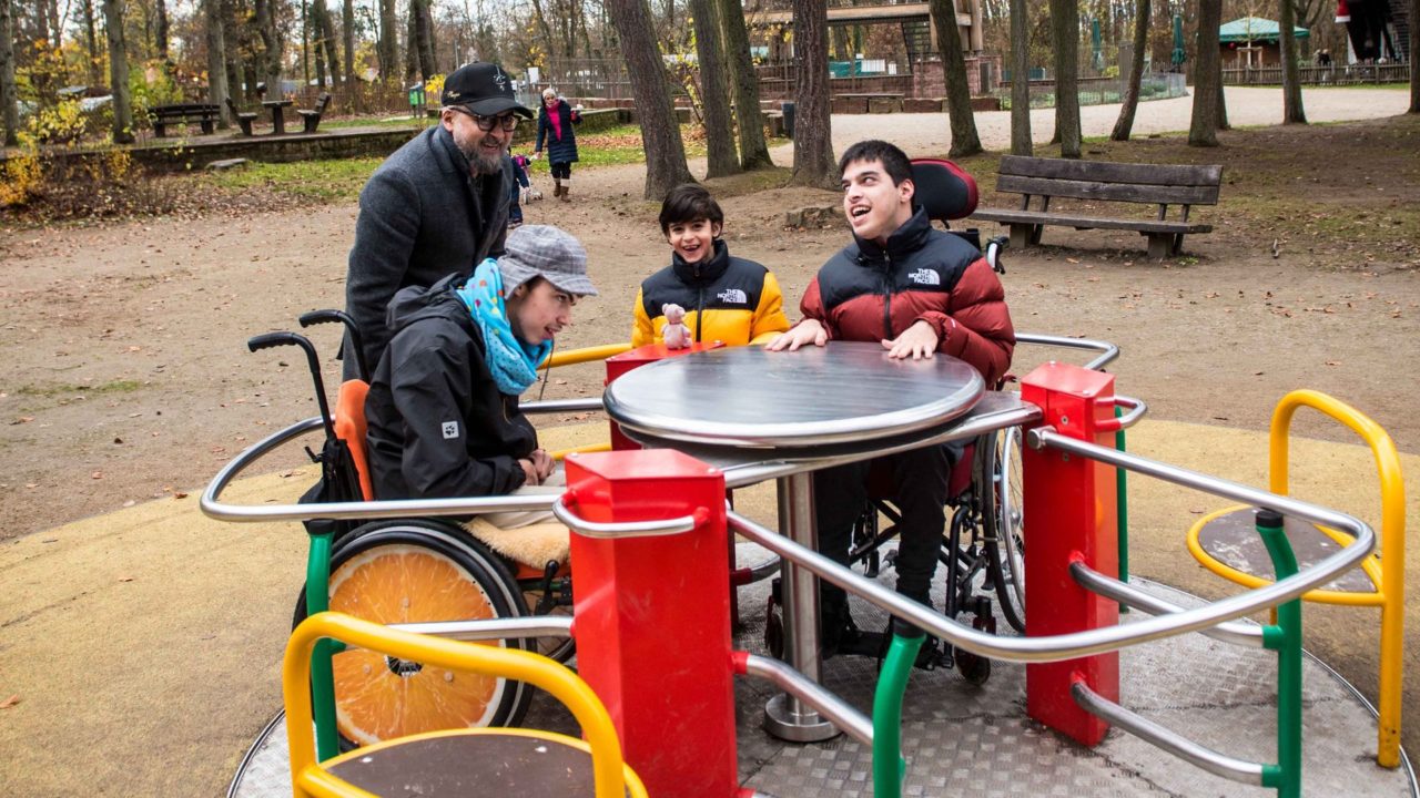 Zwei Kinder im Rollstuhl und ein Kind sitzen im neuen Rolli-Karussell auf dem Spielplatz am Goetheturm und werden von einem Vater angeschubst. Alle lachen und sind fröhlich.