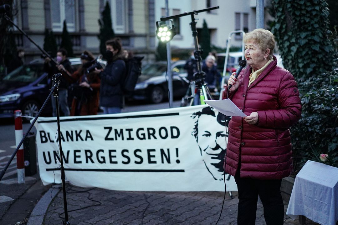 Renée Salzmann, die Tante von Blanka Zmigrod, spricht auf der Kundgebung über das Leben ihrer Tante. Sie trägt eine rote Jacke, hat kurze helle Haare und ist mit dem Banner "Blanka Zmigrod unvergessen" im Hintergrund zu sehen